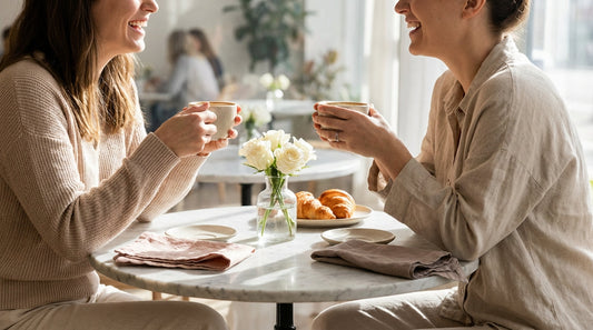 Two women laughing over coffee at a sunlit café table with white roses — Chica Bonita Coffee social ritual moment